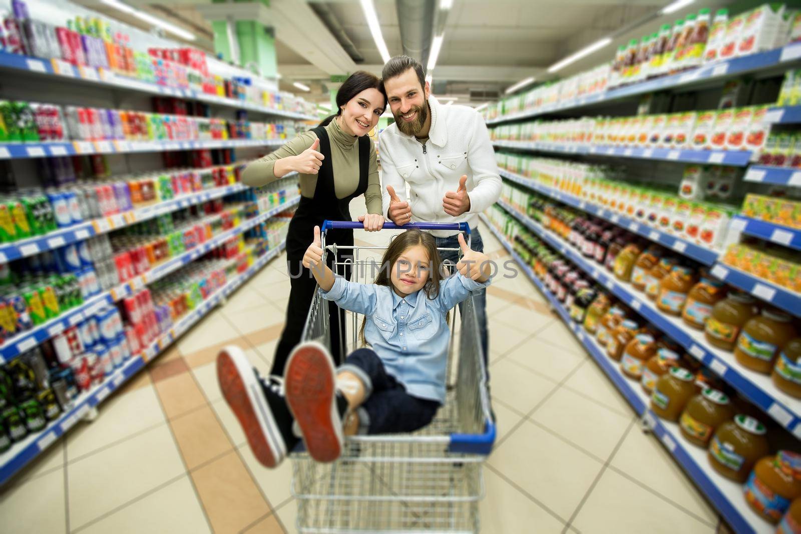 Beautiful young parents and their cute little daughter are smiling while choosing food in the supermarket by StudioPeace