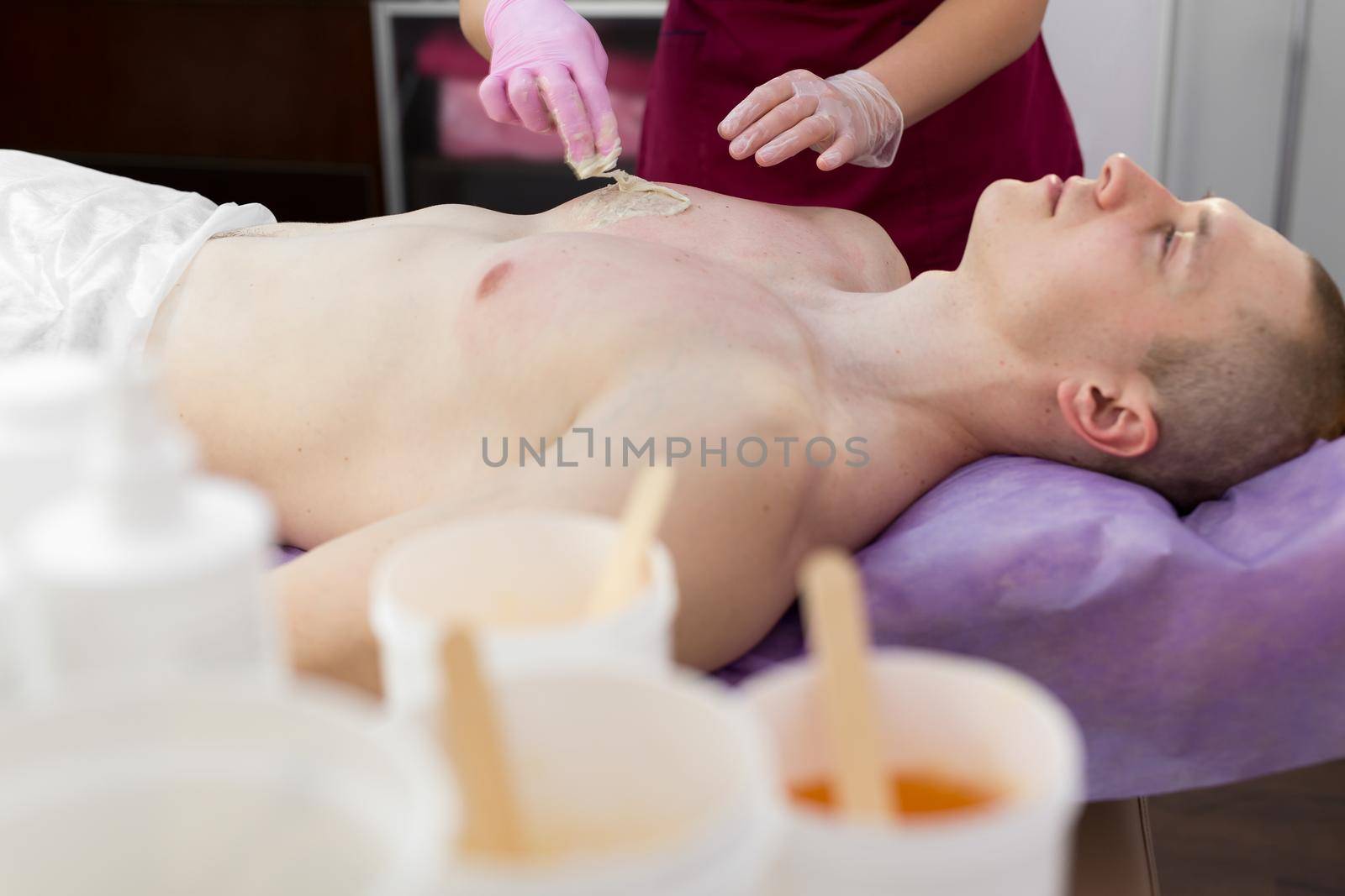 Woman beautician puts a thick sugar paste on the mans chest making him hair removal. A young guy with inflated muscles removes hair from his chest in a beauty salon. Shugaring