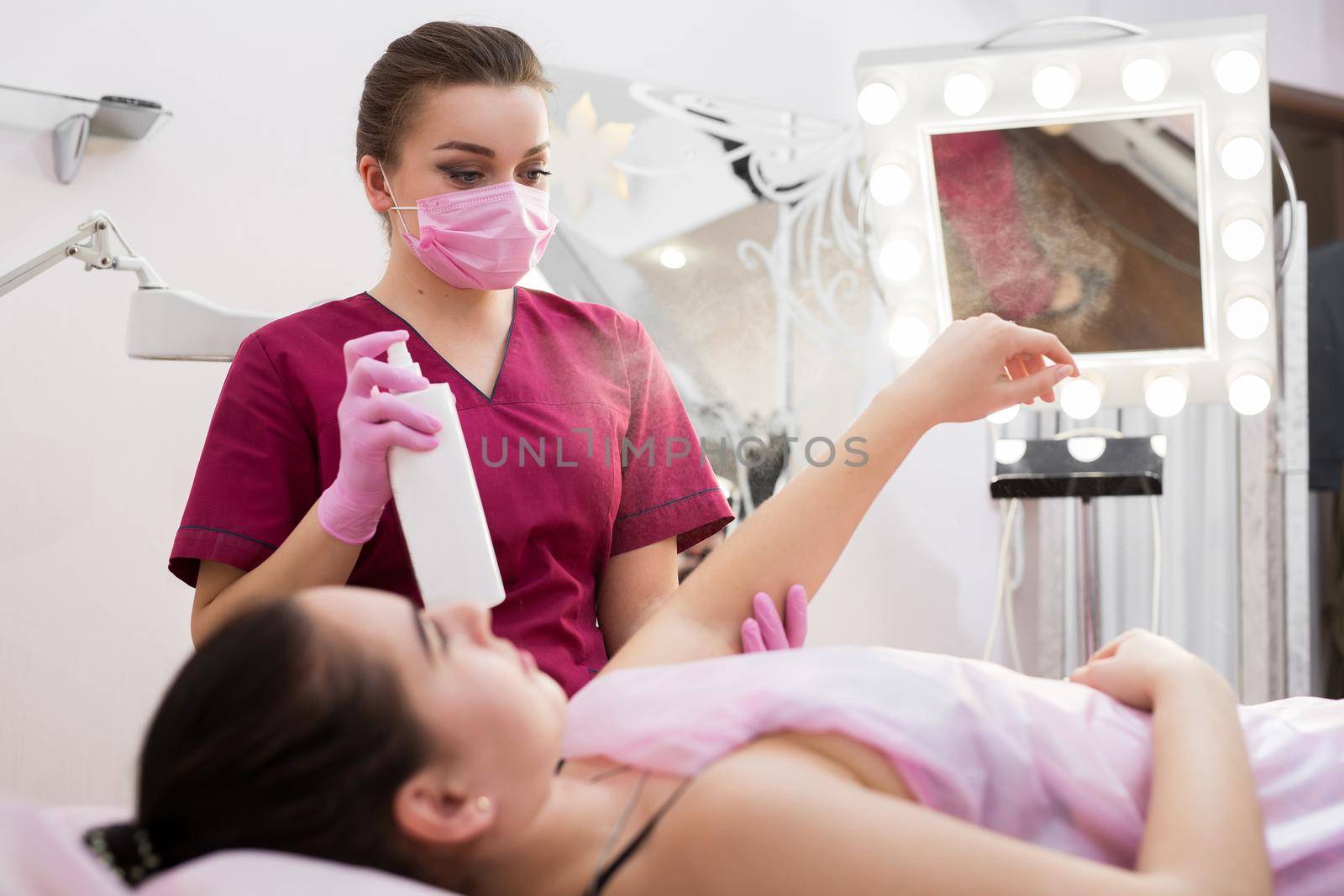 Female cosmetologist treats the hand of a young girl before the hair removal procedure. Shugaring. Master shugaring sprinkles disinfectant solution on the hand before depilation