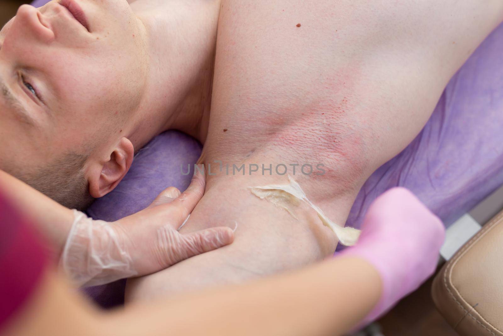 Close-up of the girl master sugaring causes a thick sugar paste on the armpits of a young sexy man. Male bodybuilder lying on the couch in the Spa salon for hair removal procedure