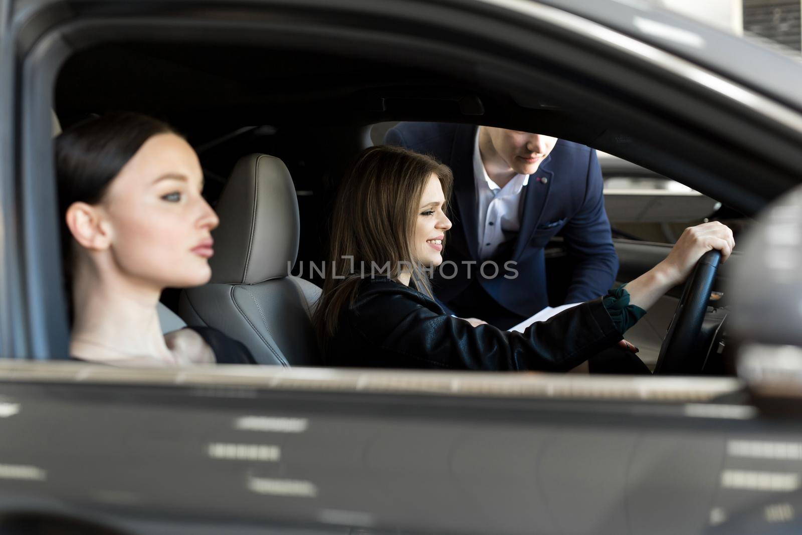 Gorgeous young girls sitting inside car, smiling and looking at camera. One of girls holding hands on steering wheel. Female customers of car dealership choosing auto in showroom by StudioPeace