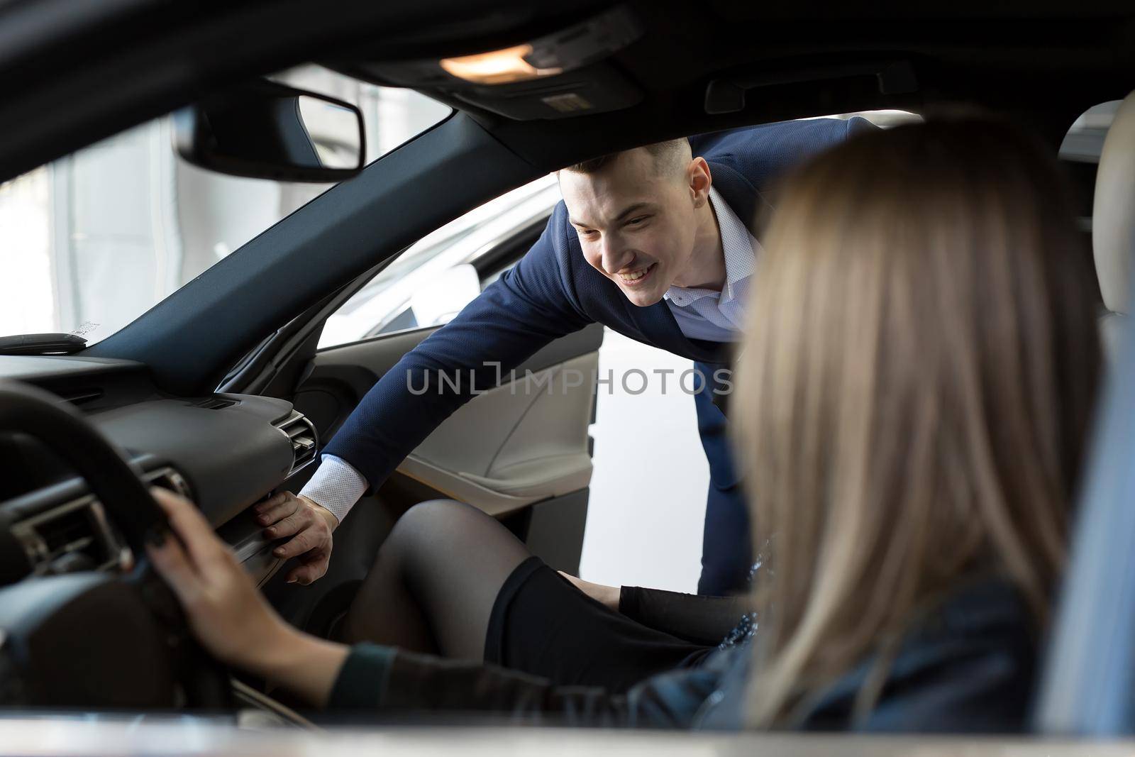 Side view of young beautiful woman sitting inside car and holding hand on steering wheel. She smiling and talking with manager of car dealership. Car agent representing inside of automobile.