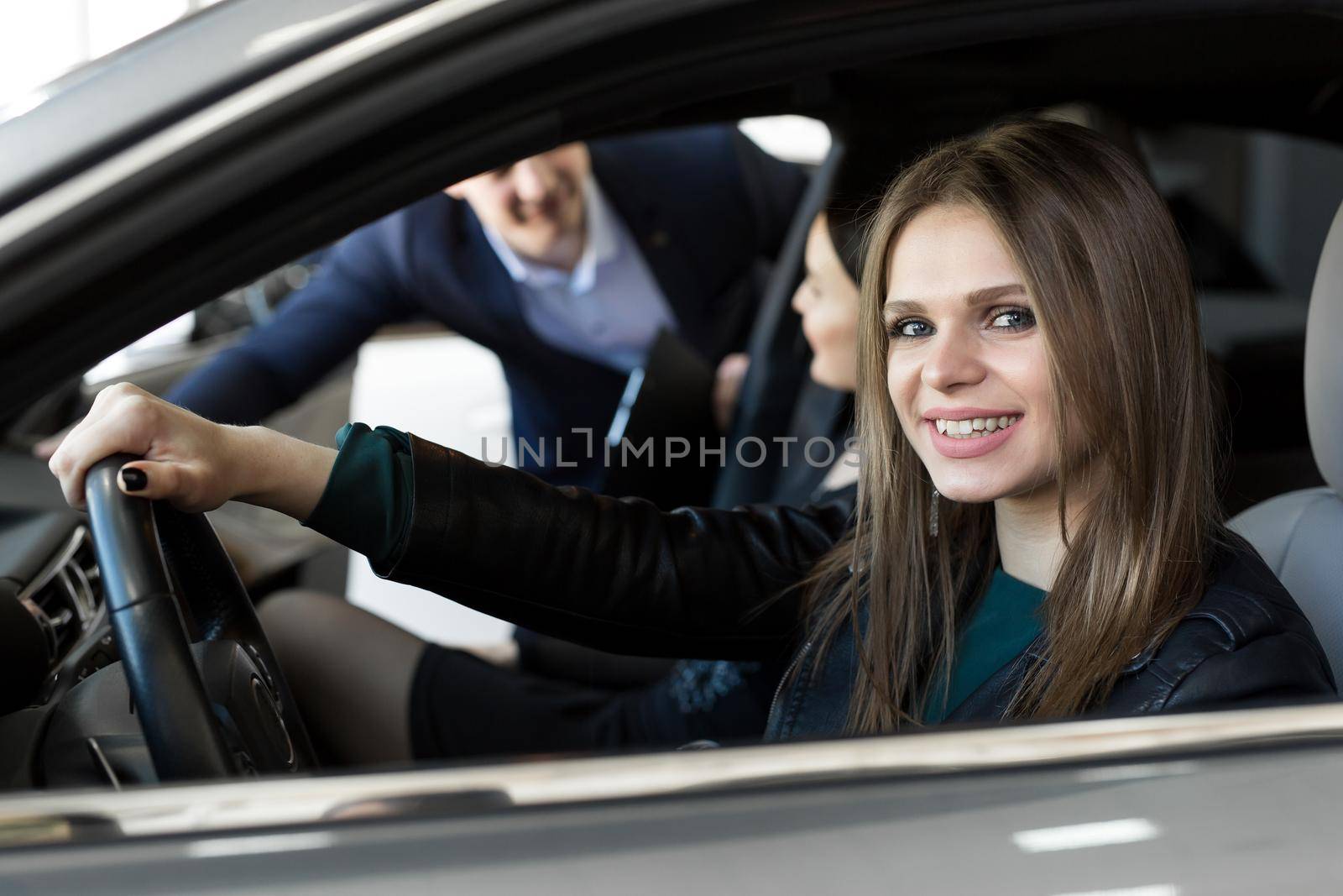 Side view of young beautiful woman sitting inside car and holding hand on steering wheel. She smiling and talking with manager of car dealership. Car agent representing inside of automobile.