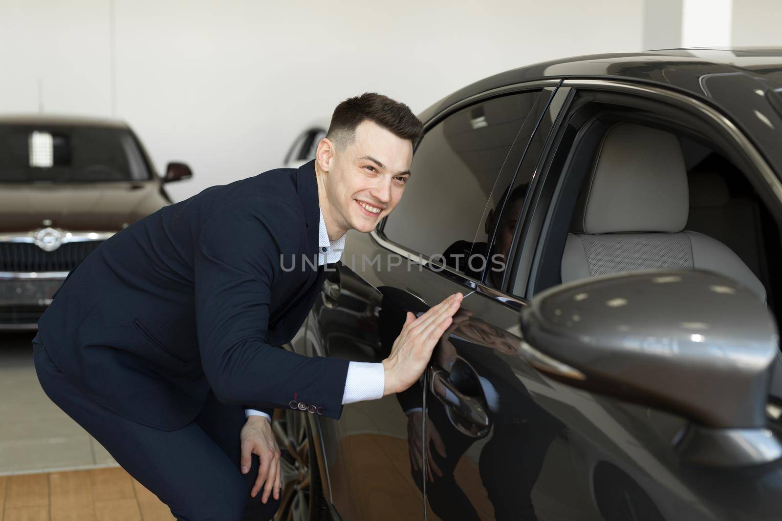 Handsome young businessman in classic blue suit is smiling while examining car in a motor show. by StudioPeace