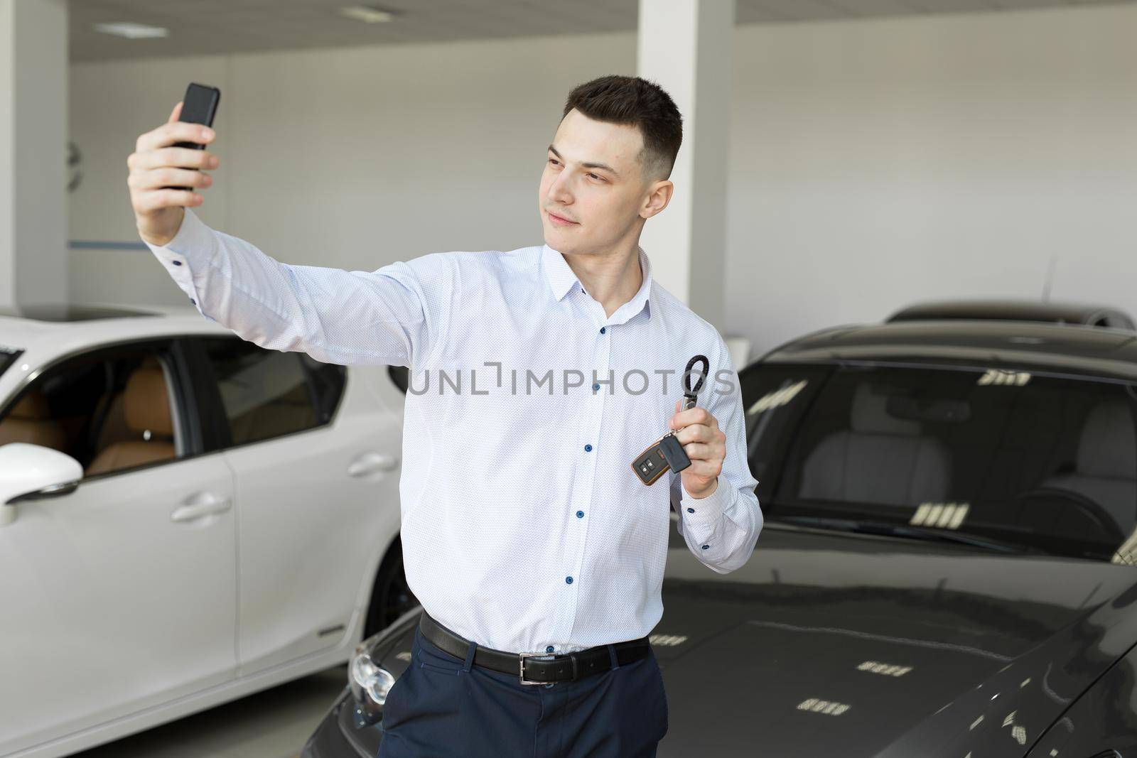 Happy businessman making selfie photo holding keys in front of his new car in the showroom. by StudioPeace