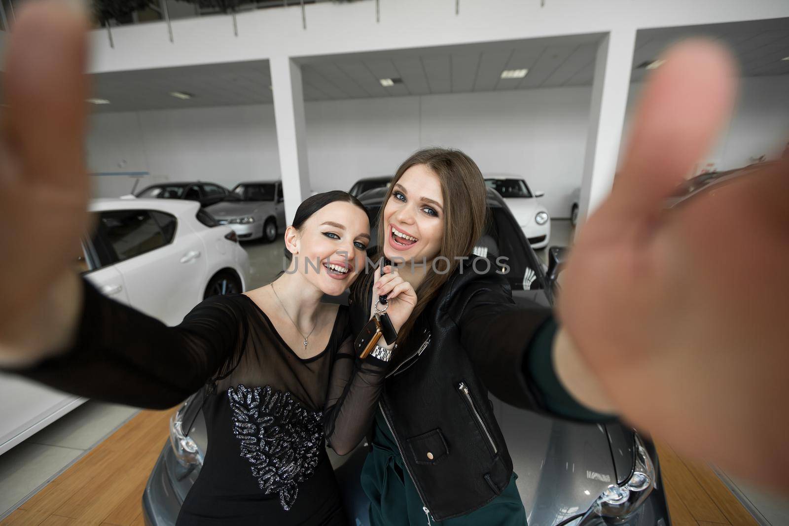 Beautiful and cute girls making selfie near red car. Young female customers smiling and hugging. Girls happy because of buying new automobile in modern car dealership by StudioPeace
