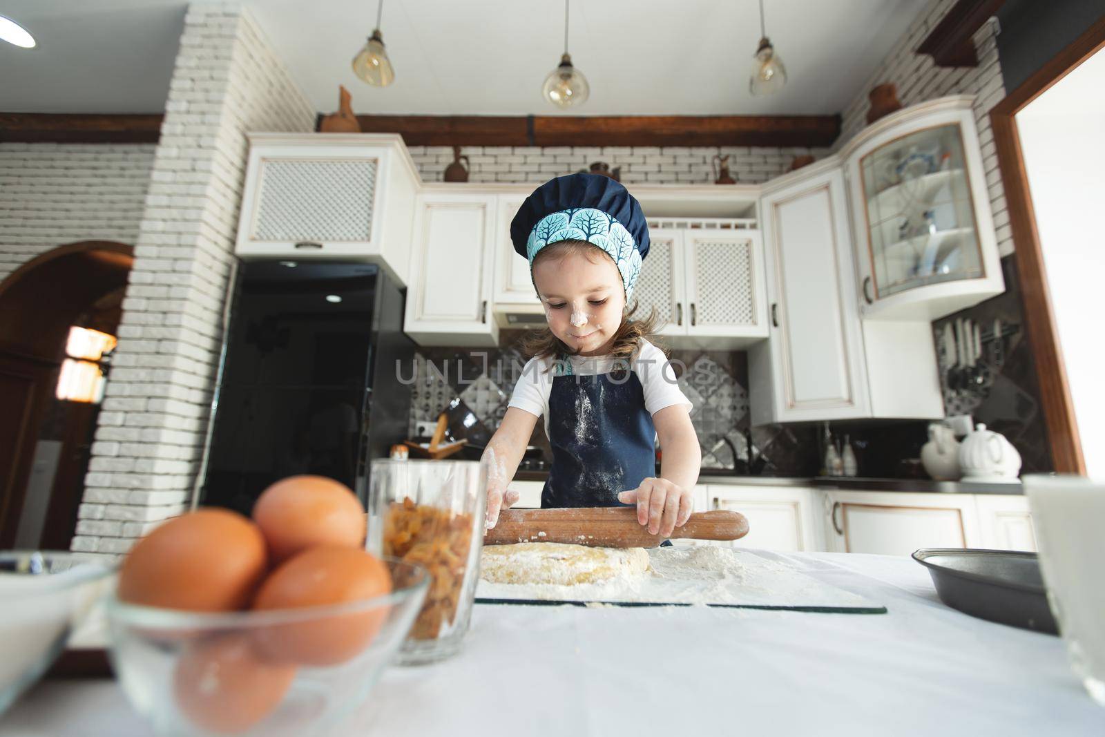 A little girl in an apron and a chef's hat is rolling out cookie dough, laughing.