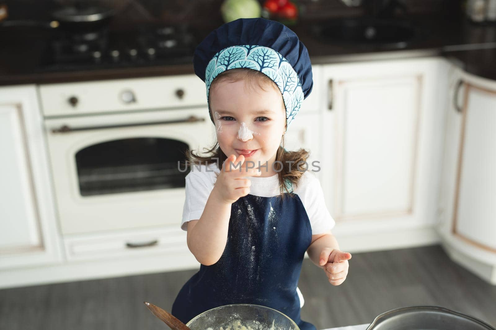 Cute little girl in apron and chef hat is flattening the dough using a rolling pin, looking at camera and smiling while baking