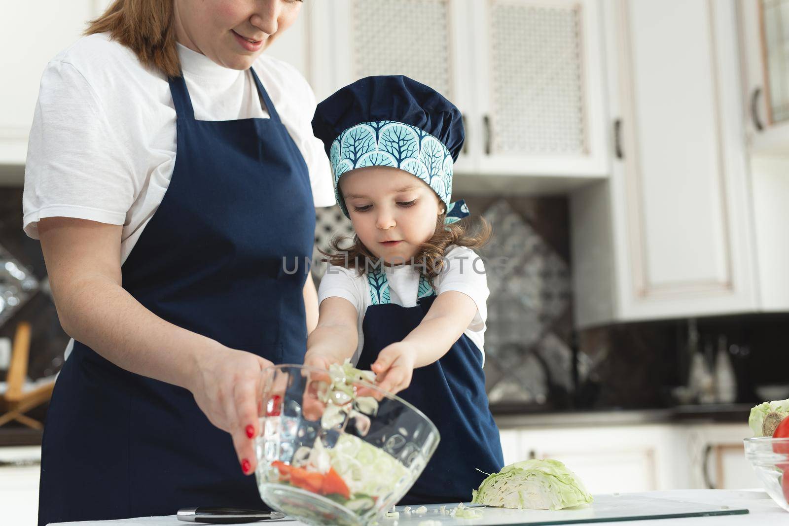 Caring mother teaching little daughter to cook salad in kitchen, young mum and adorable cute girl child wearing apron chopping vegetables with knife on countertop, standing in kitchen at home