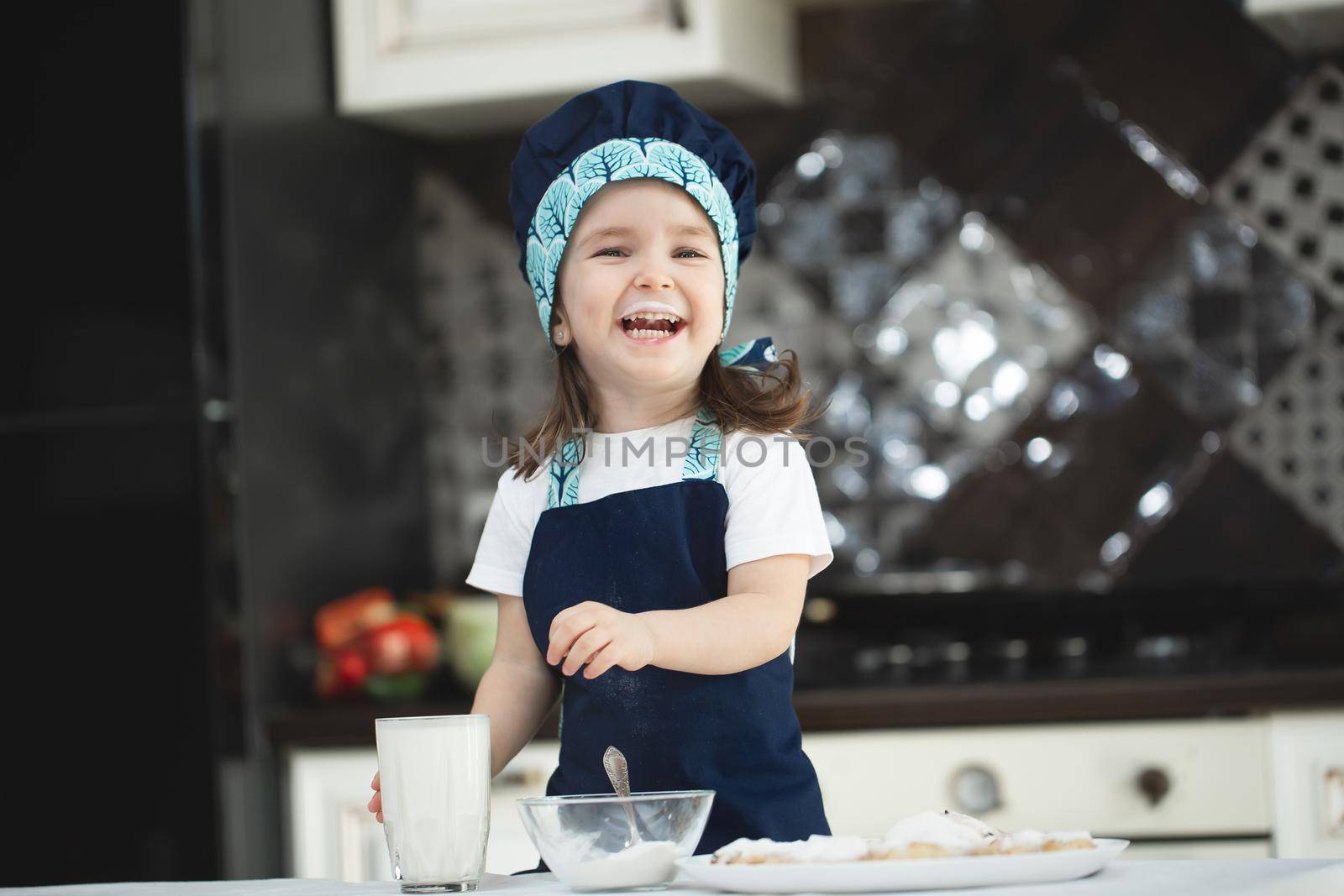 Small girl in the kitchen in an apron and a Chef's hat is drinking milk from a glass glass.