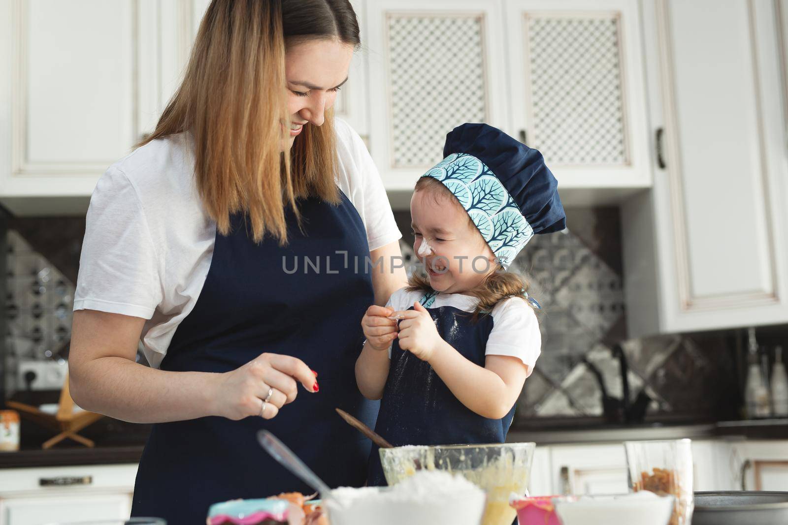Cute little girl and her beautiful mom in matching aprons and caps play and laugh while kneading dough in the kitchen by StudioPeace