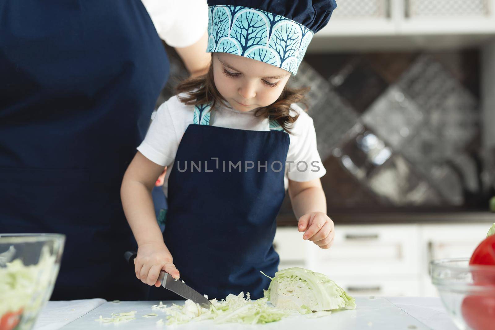 Caring mother teaching little daughter to cook salad in kitchen, young mum and adorable cute girl child wearing apron chopping vegetables with knife on countertop, standing in kitchen at home. by StudioPeace