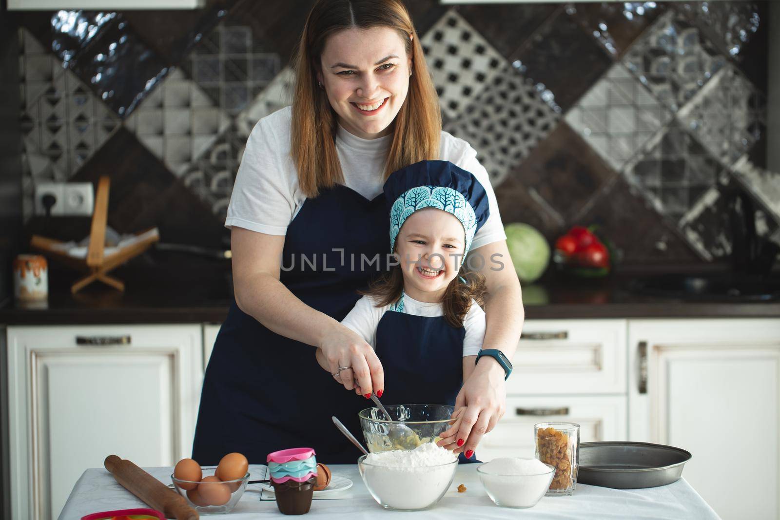 Mother and daughter preparing a sweet cake using flour, milk, sitting on chairs at a table in a modern kitchen. Girl holding a whisk, stirring eggs in a bowl, preparing pancake dough with her mom.