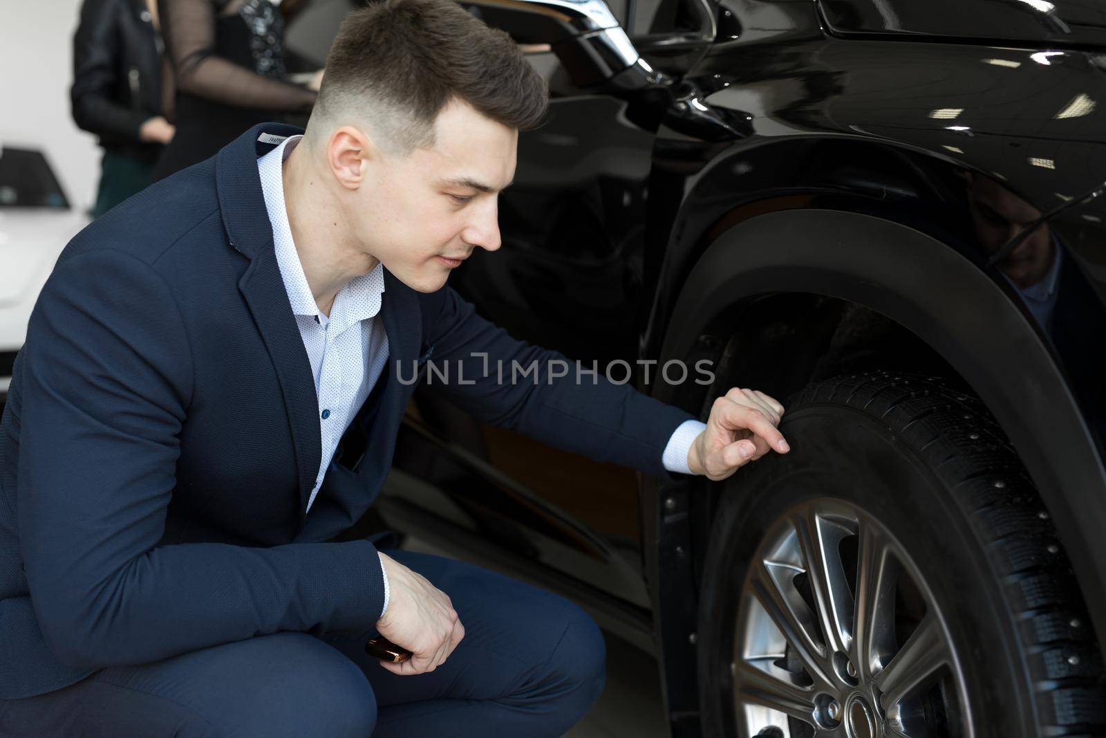 Attractive elegant man examining wheels of a new automobile on sale at dealership. Handsome male driver choosing new car to buy