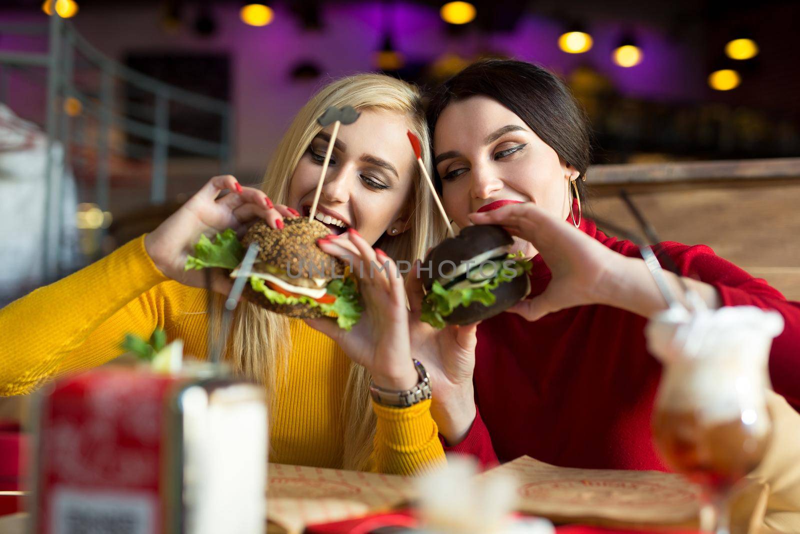 Two happy girls bite burgers. Concept of fast food. by StudioPeace