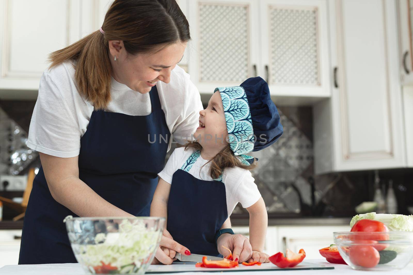 Caring mother teaches her little daughter how to prepare a salad in the kitchen, a young mother and a charming sweet baby girl look at each other and smile by StudioPeace