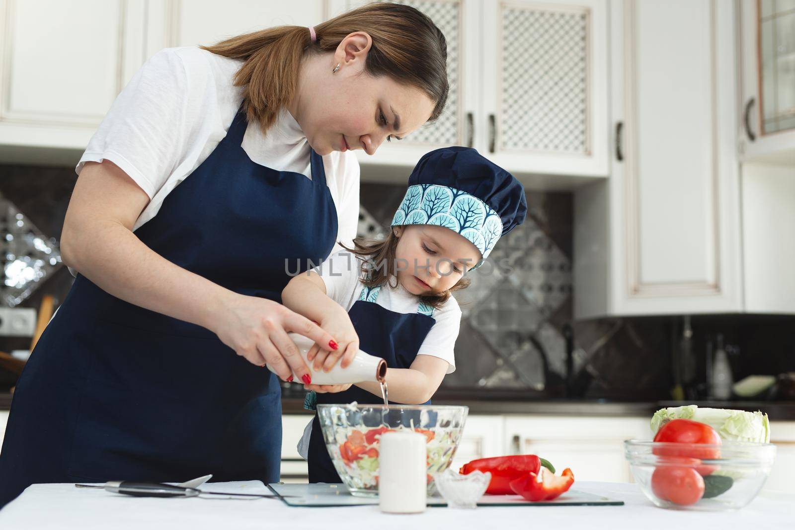 A caring mother teaches her little daughter how to prepare a salad in the kitchen, a young mother and a charming sweet baby girl dress up the salad, salt it and taste it.