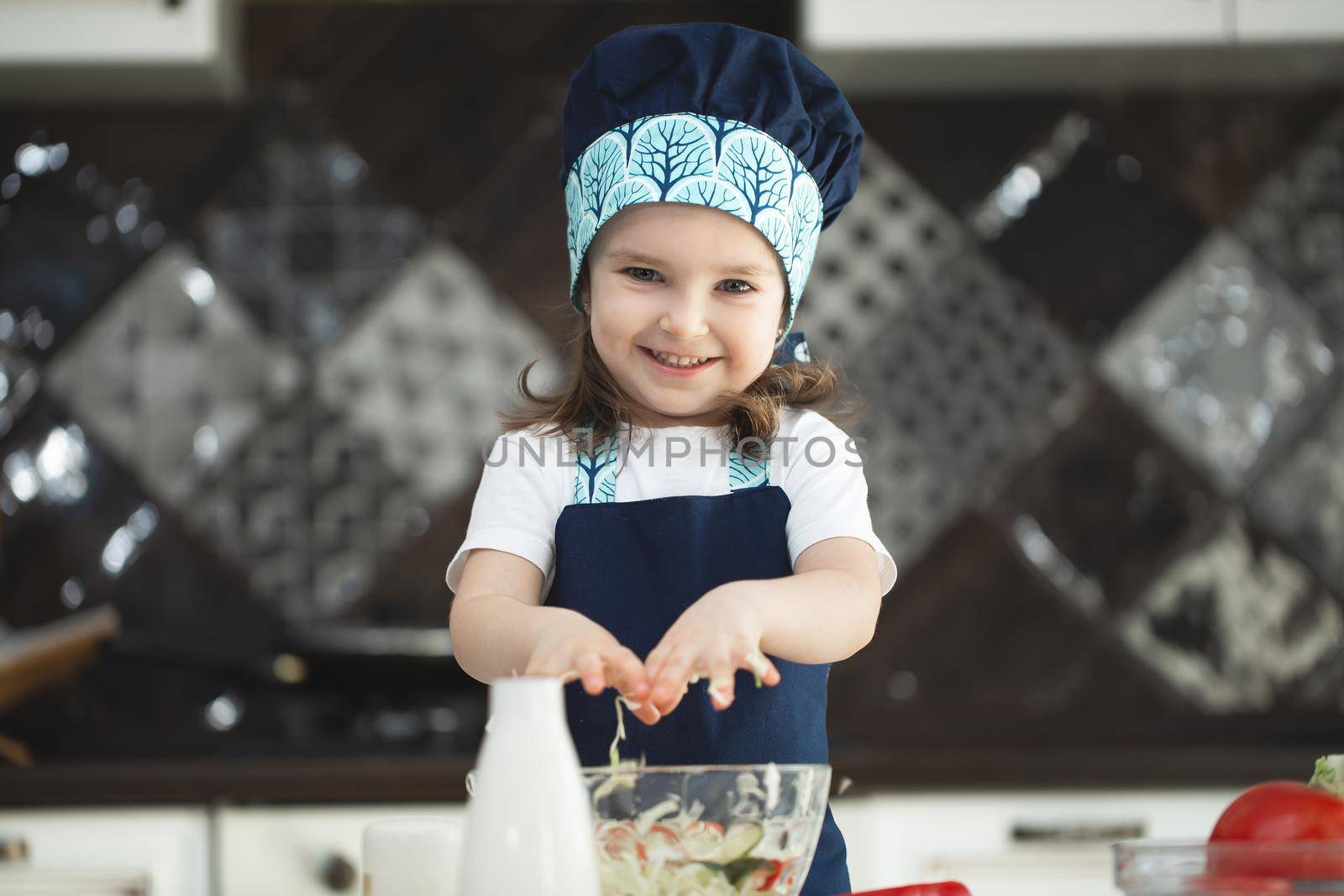 A child in an apron and a Chef's hat is stirring a vegetable salad in the kitchen and looking at the camera by StudioPeace