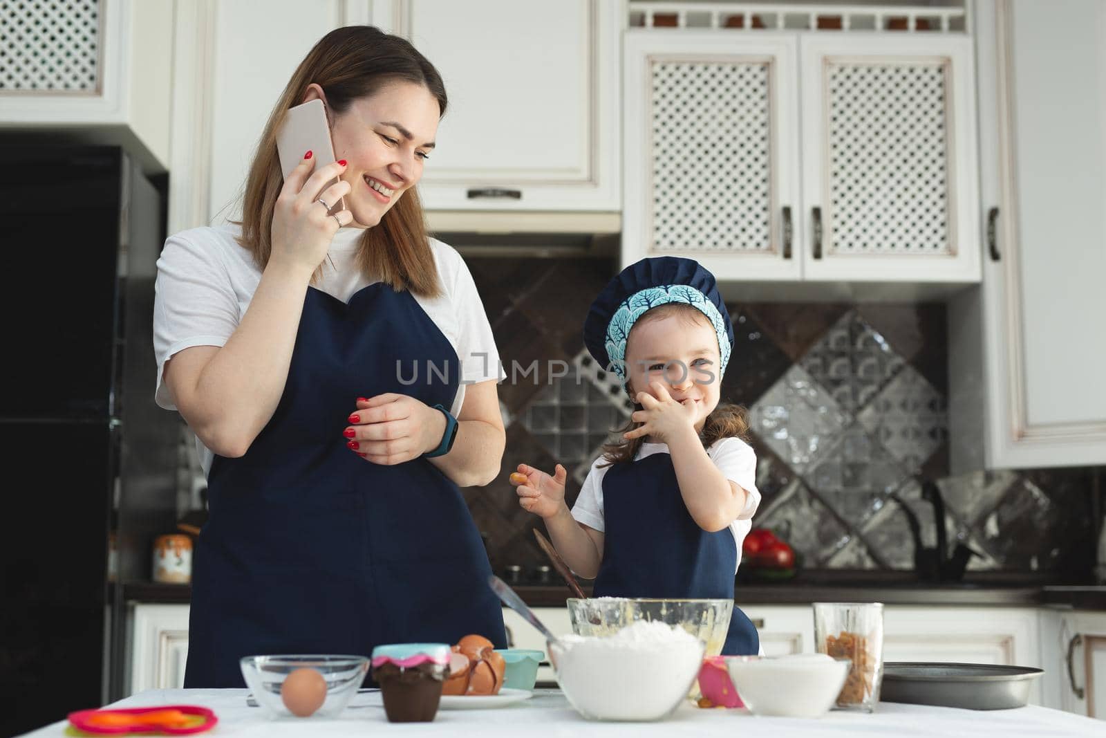 A mother and daughter in identical aprons and chef's hats are cooking in the kitchen, smiling and looking at the camera. Mom's on the phone by StudioPeace