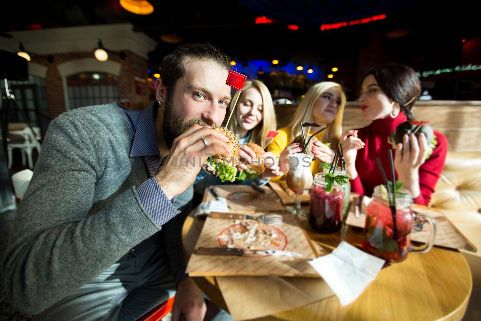 The man takes a bite of his Burger and looking at the camera. Beautiful women laugh and communicate.