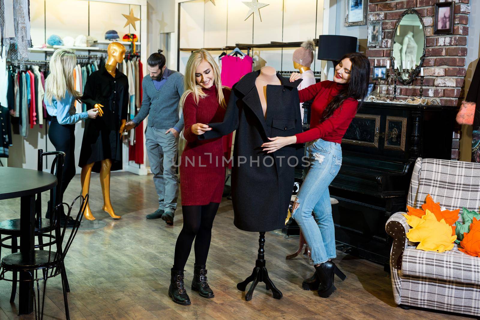 Shopping, fashion, clothes, style and people concept - happy friends trying coat on in mall or clothing store. by StudioPeace