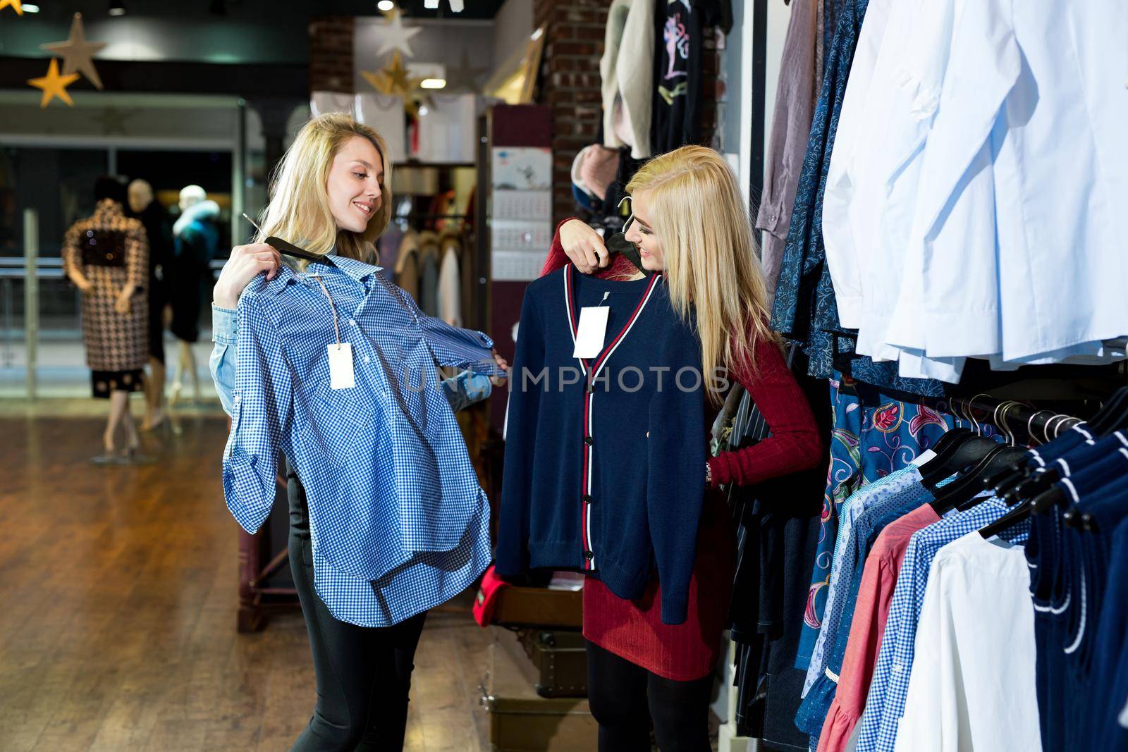 Two happy female best friends looking at the quality of a shirt hanging on a rail inside the clothing store. by StudioPeace