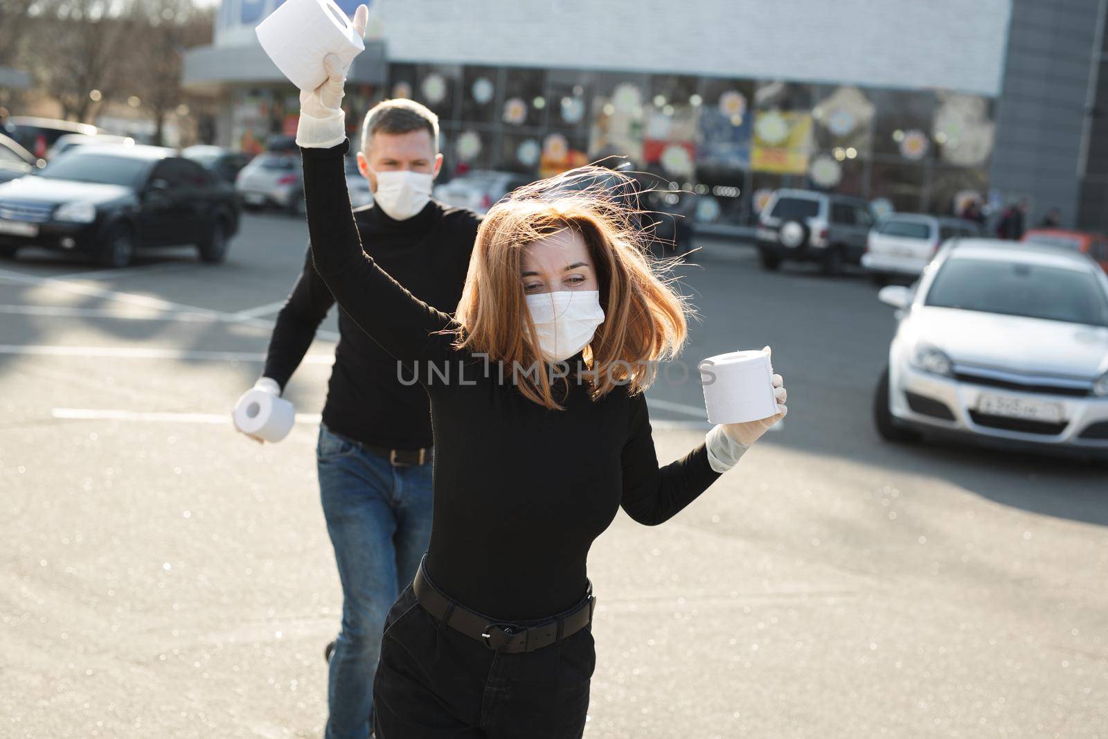 Woman and a man in a coronavirus face mask hold large rolls of toilet paper on a city street and indulge by StudioPeace