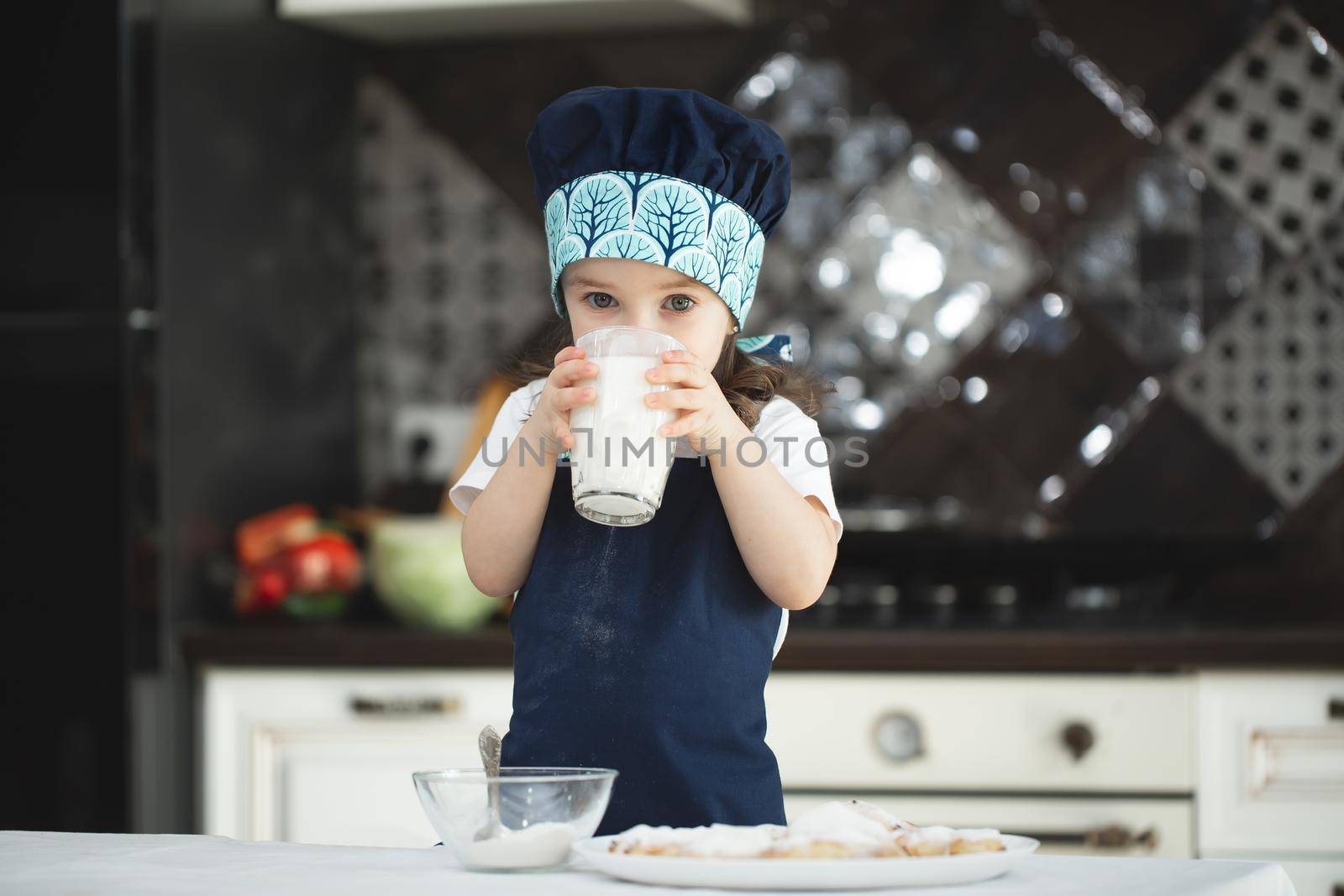 Small girl in the kitchen in an apron and a Chef's hat is drinking milk from a glass glass.