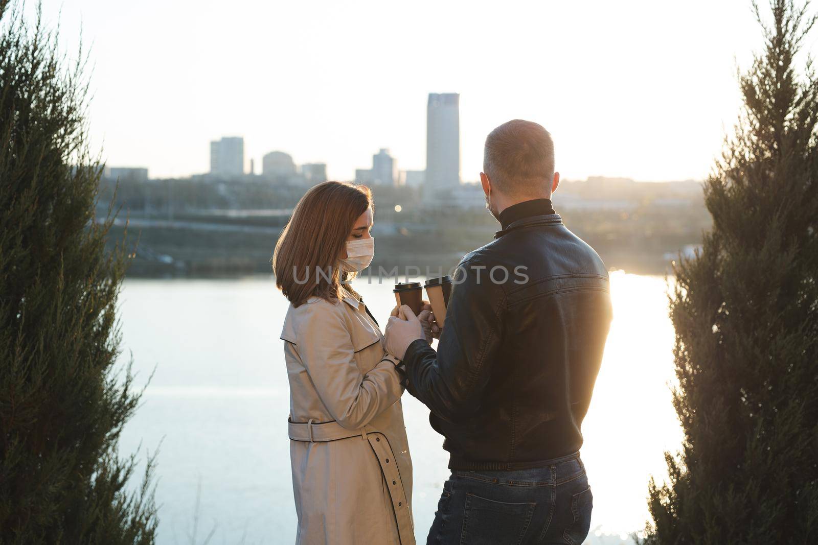 Young couple, a man and a woman in medical masks and gloves, drink coffee from disposable cups on the street and look at each other against the background of the river and the city. Quarantine.