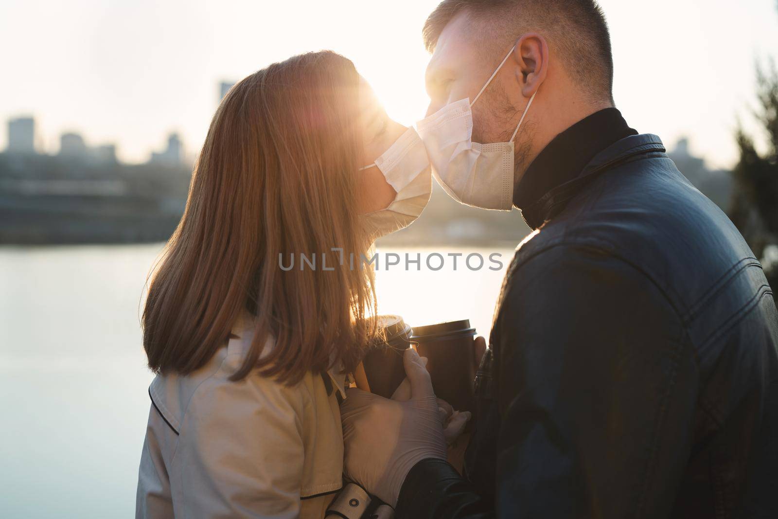 Young couple, a man and a woman in medical masks and gloves, drink coffee from disposable cups on the street and kiss. Coronavirus. Covid 19. by StudioPeace