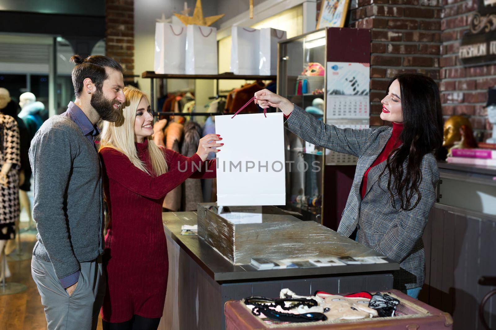 Attractive saleswoman, handing over a bag of clothes to a beautiful couple of men and women. by StudioPeace