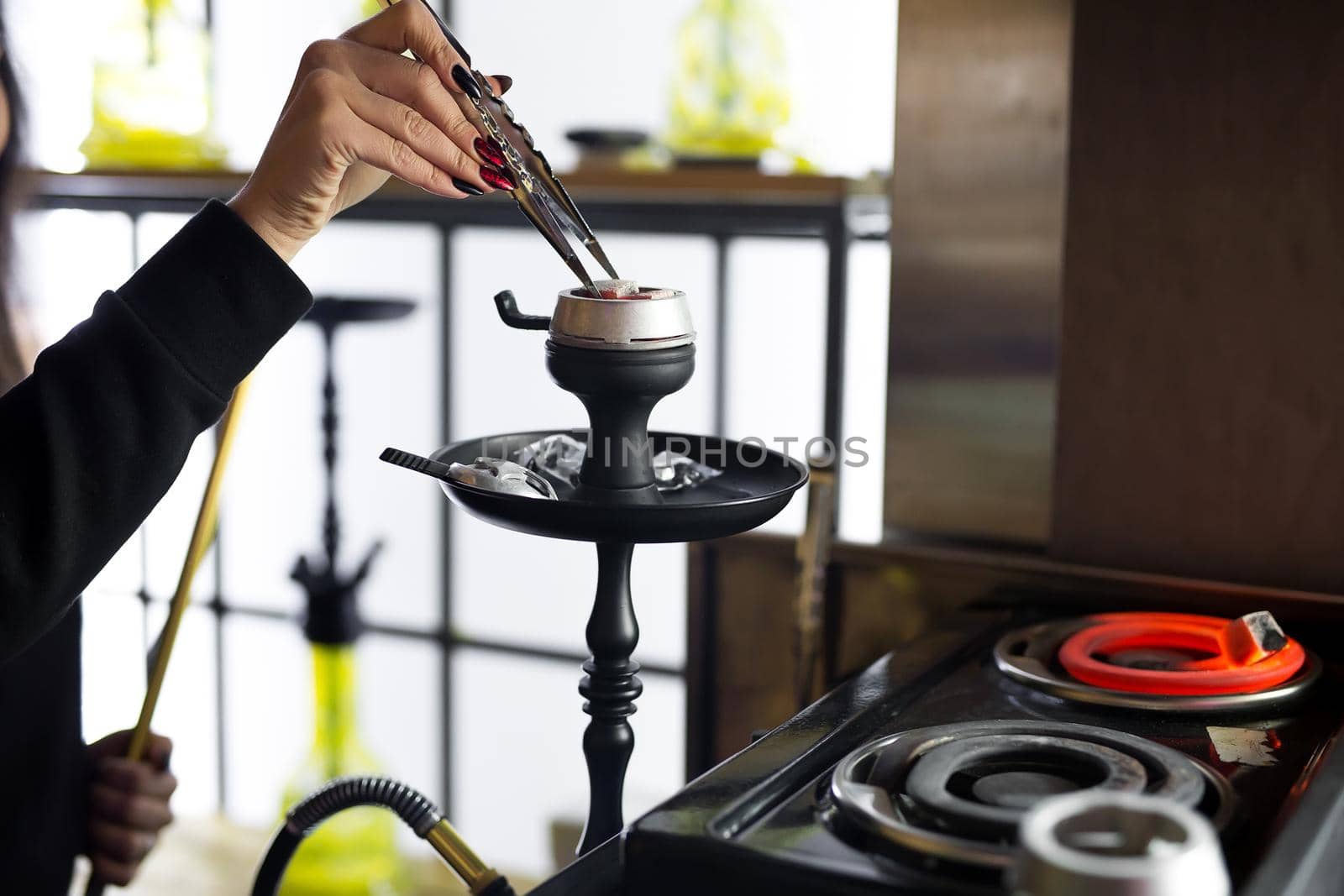 Women's hands hold hookah tongs and adjust the hot coals in a metal bowl. Black hookah stands in a restaurant or bar by StudioPeace