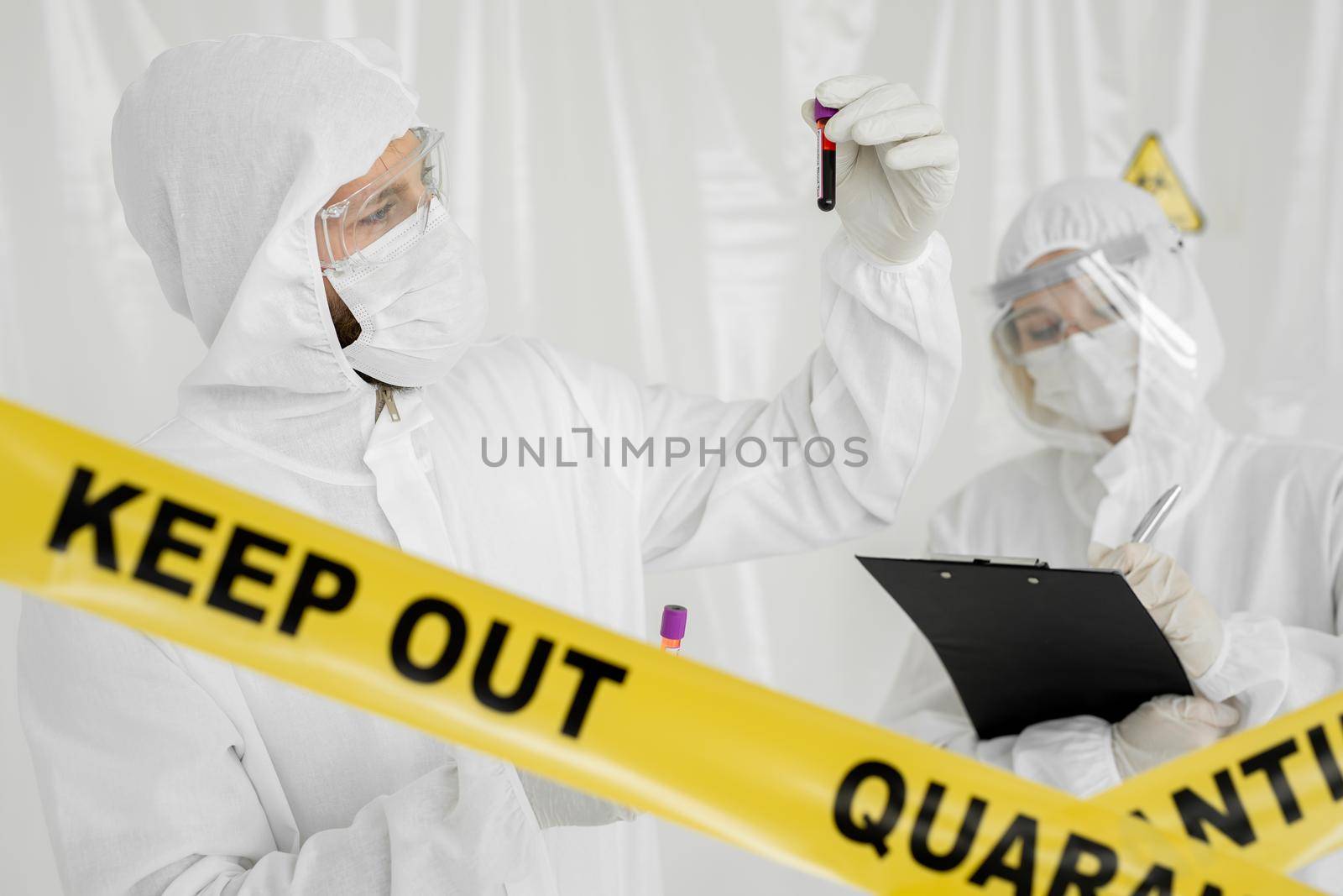 Epidemiologist woman in protective clothing is in a restricted area with a planchette. Infected blood sample in sample tube in hand of scientist doctor biohazard protection clothing.