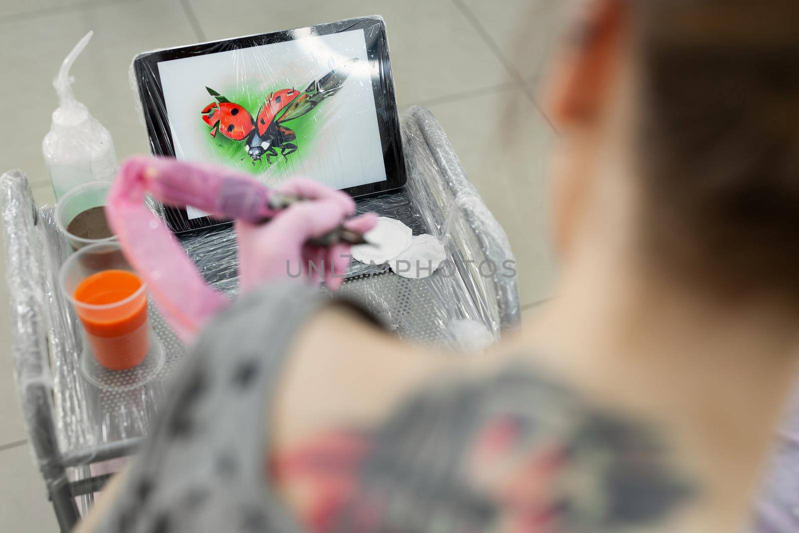 Tattoo master dips a tattoo machine with needles in black ink. The tattoo master applies a colored tattoo on the skin of a young girl, in the background is a tablet with a picture of a ladybug