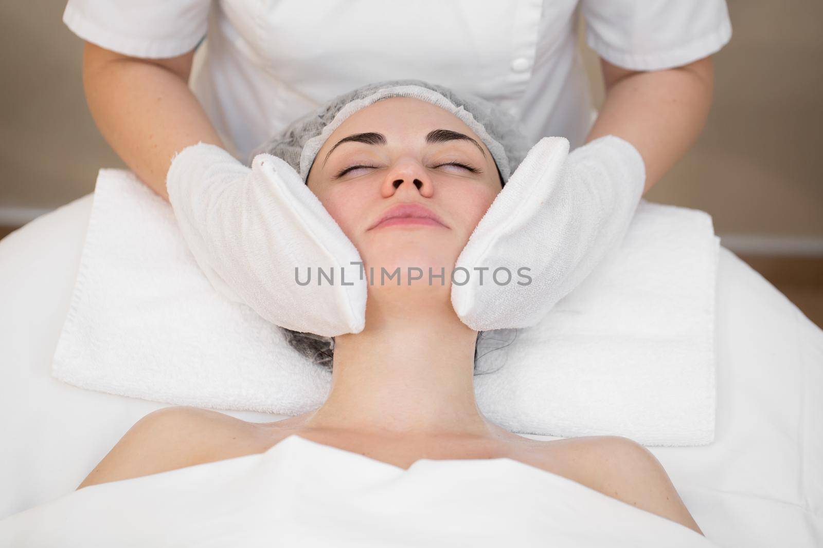 Hands of the beautician wipe the girl's face in the cosmetology center with white gloves. Portrait of a beautiful girl on the procedure for rejuvenating the face in a beauty salon. by StudioPeace