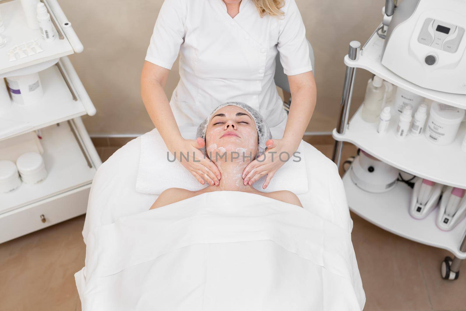 Beautiful girl at the beautician does the spa procedures, facial massage and mask for the skin. The hands of a beautician do a facial massage with a moisturizing cream to a young girl in the spa salon. by StudioPeace
