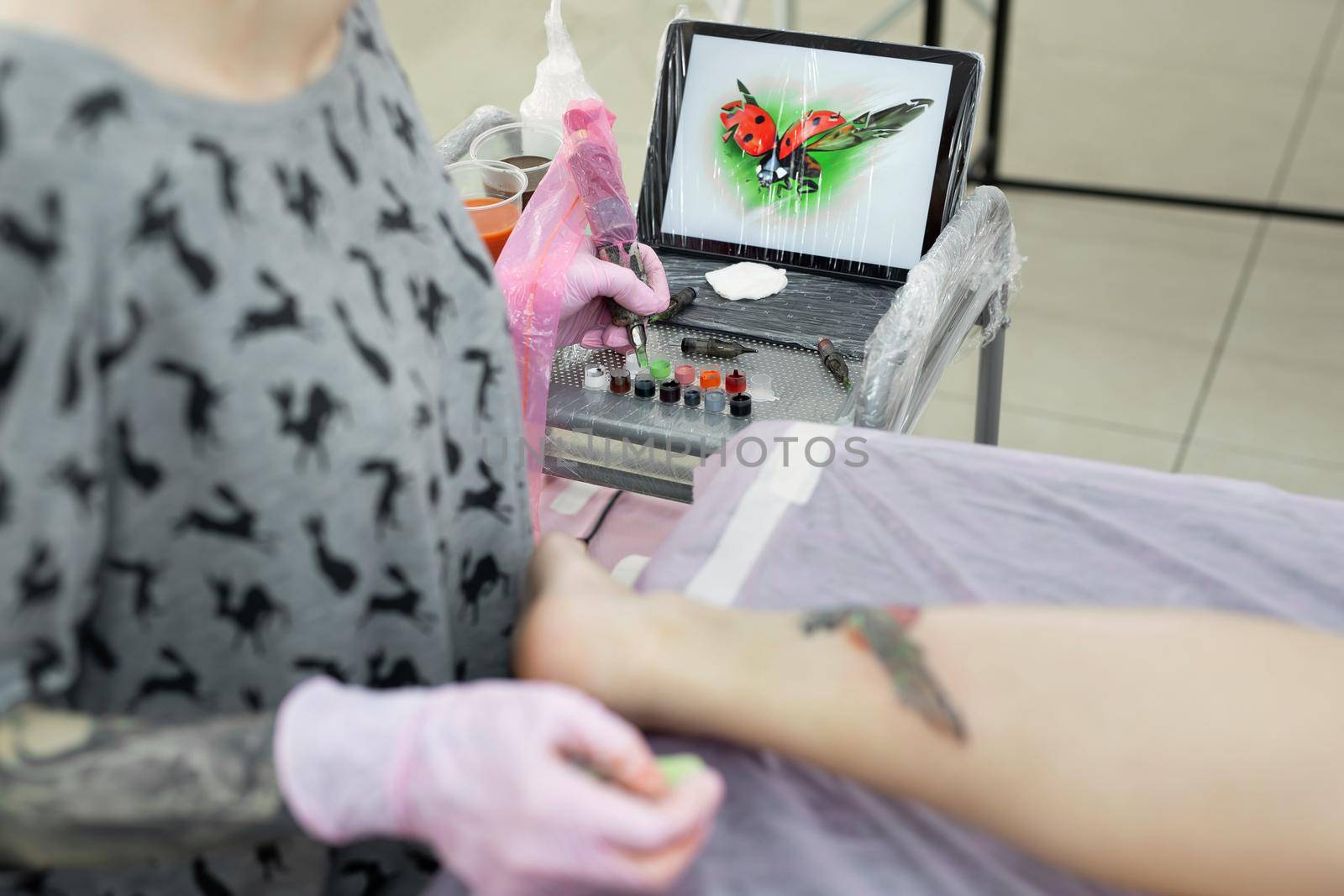 Tattoo master dips a tattoo machine with needles in black ink. The tattoo master applies a colored tattoo on the skin of a young girl, in the background is a tablet with a picture of a ladybug. by StudioPeace
