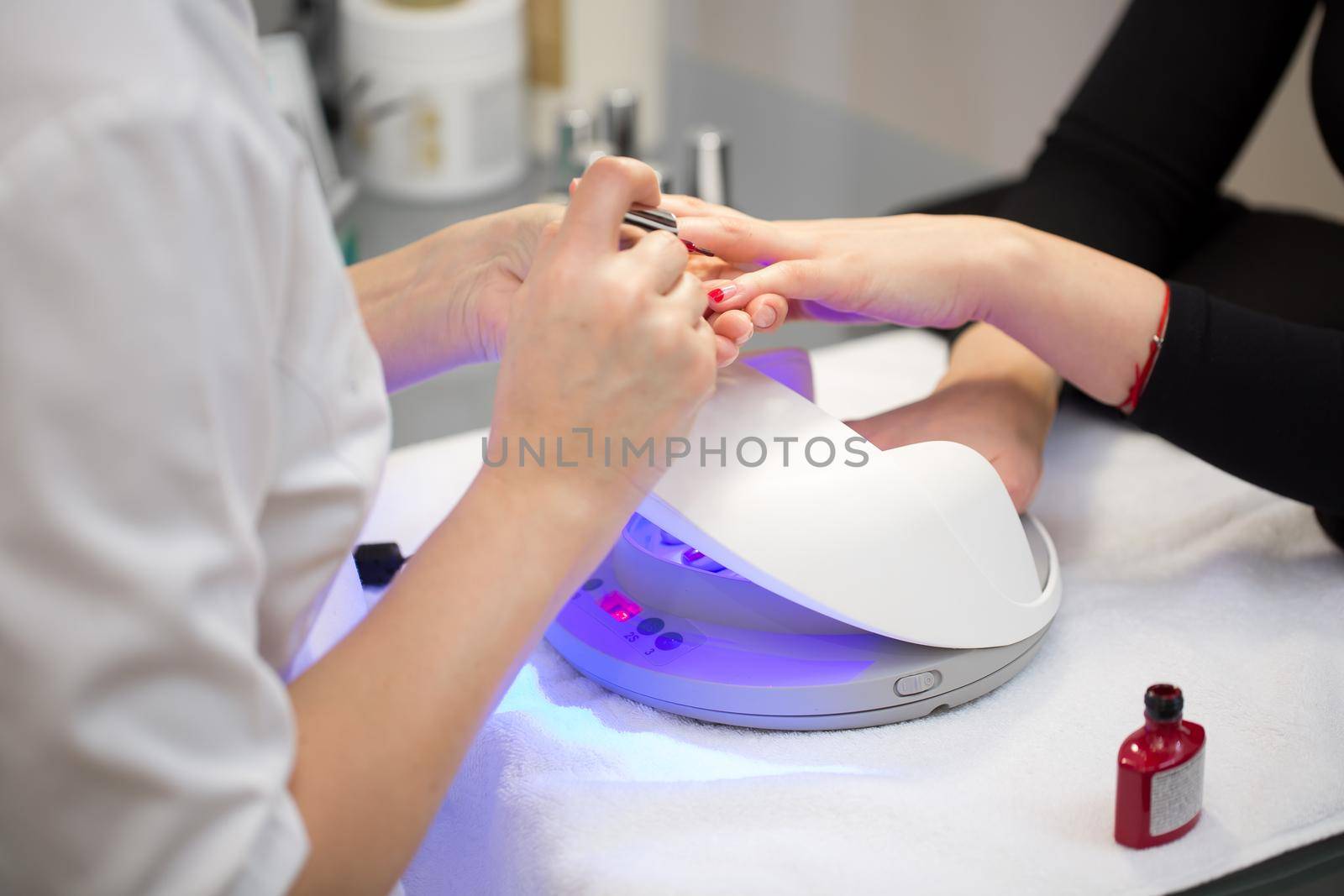 Close-up of the hand of a manicurist who applies red gel Polish on the nails of a young girl. Girl dries gel nail Polish in UV lamp by StudioPeace