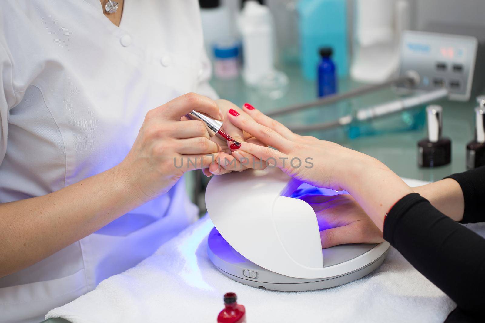Close-up of the hand of a manicurist who applies red gel Polish on the nails of a young girl. Girl dries gel nail Polish in UV lamp.