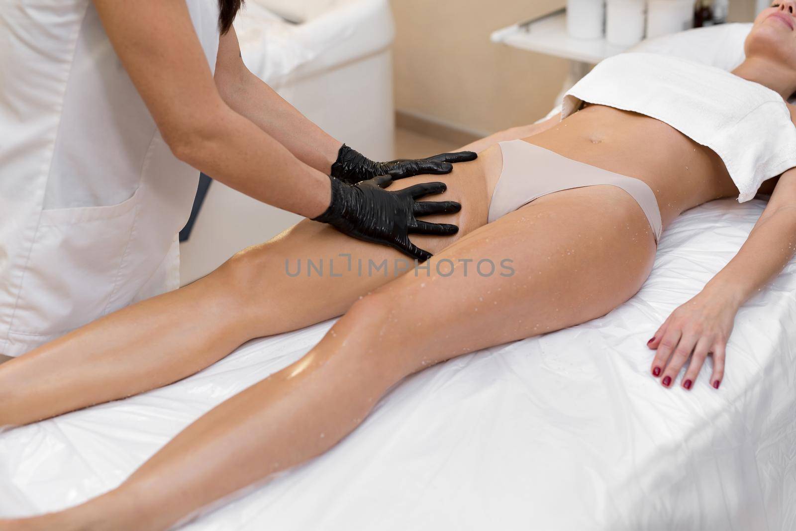 Young girl beautician does a massage to a woman, she rubs her legs with oil. Anti-cellulite massage in the spa salon.