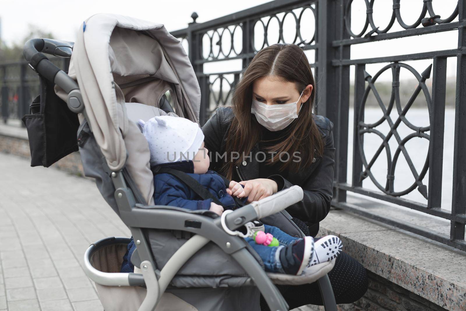Woman with a mask on her face to protect against coronavirus, covid-19 holds her young child by the hand, who is sitting in a stroller