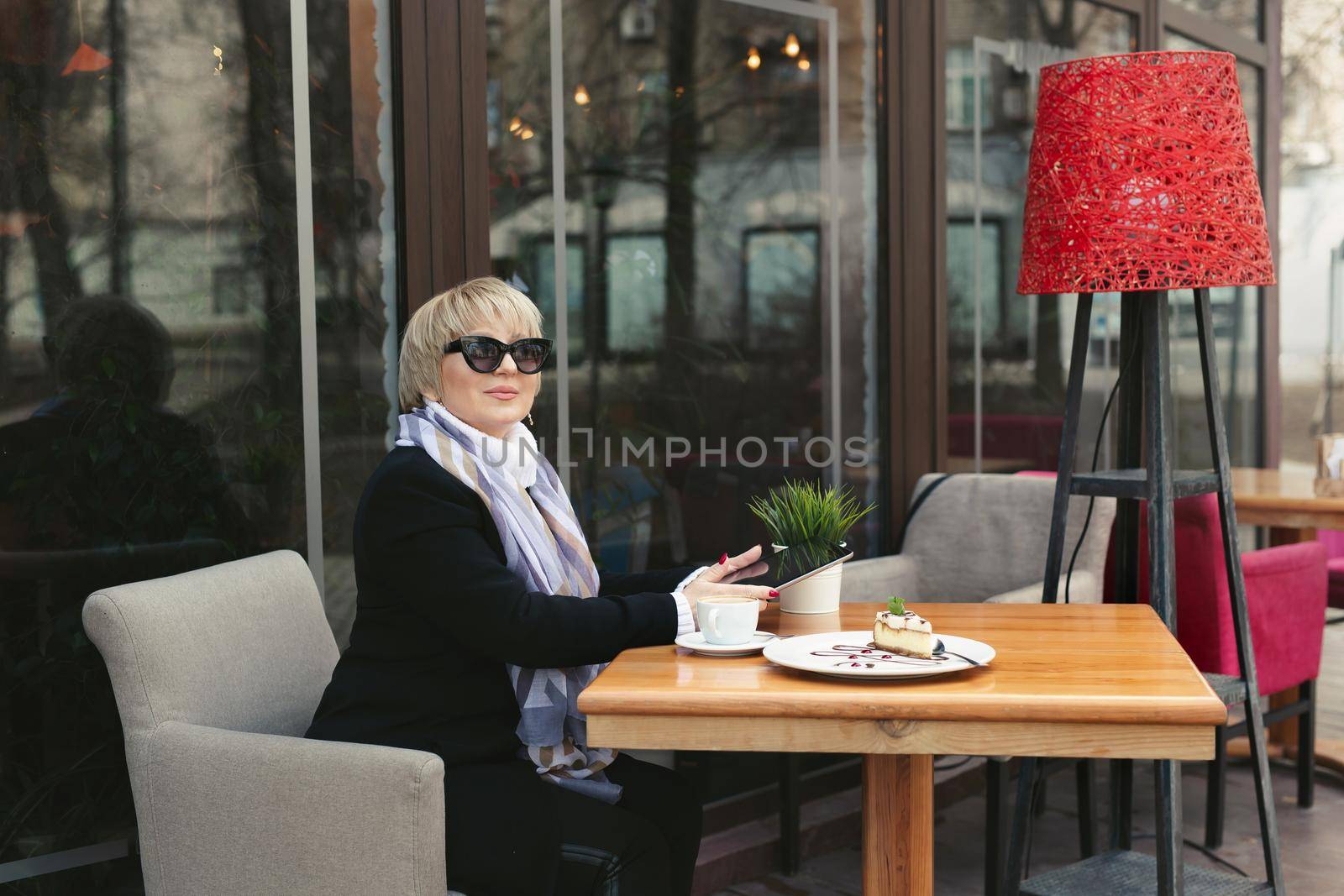 An elderly woman in black glasses works on a tablet in a cafe on the street during lunch.