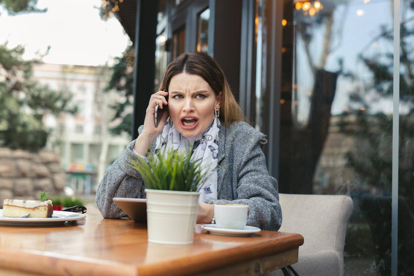 Portrait of angry young woman solving business problems shouting during phone conversation sitting in cafe terrace.Disappointed female upset about getting bad news from employee talking on cellular by StudioPeace