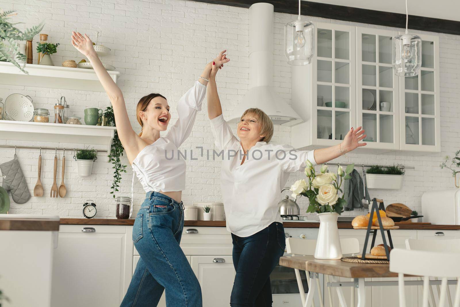 Funny elderly mother is having fun in the kitchen with an adult daughter, they cook dinner and sing together while holding kitchen utensils.