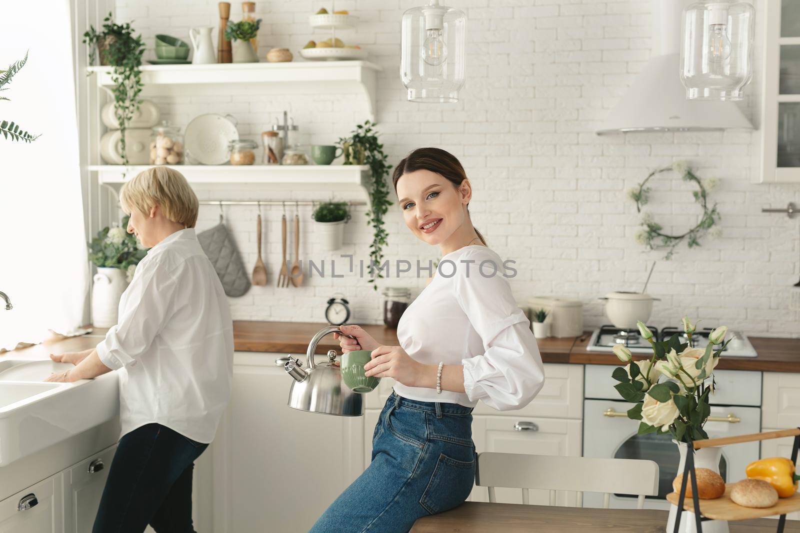 Adult daughter pours tea from a teapot into a mug while her elderly mother washes dishes in the kitchen sink.