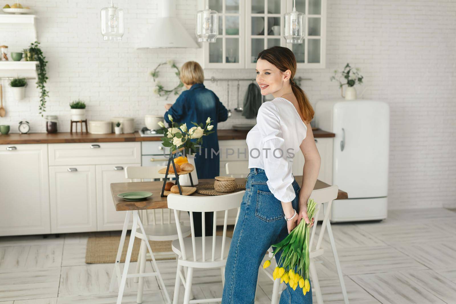 Beautiful adult daughter hides flowers behind her back for her adult mother. Mother's day, a women's holiday.