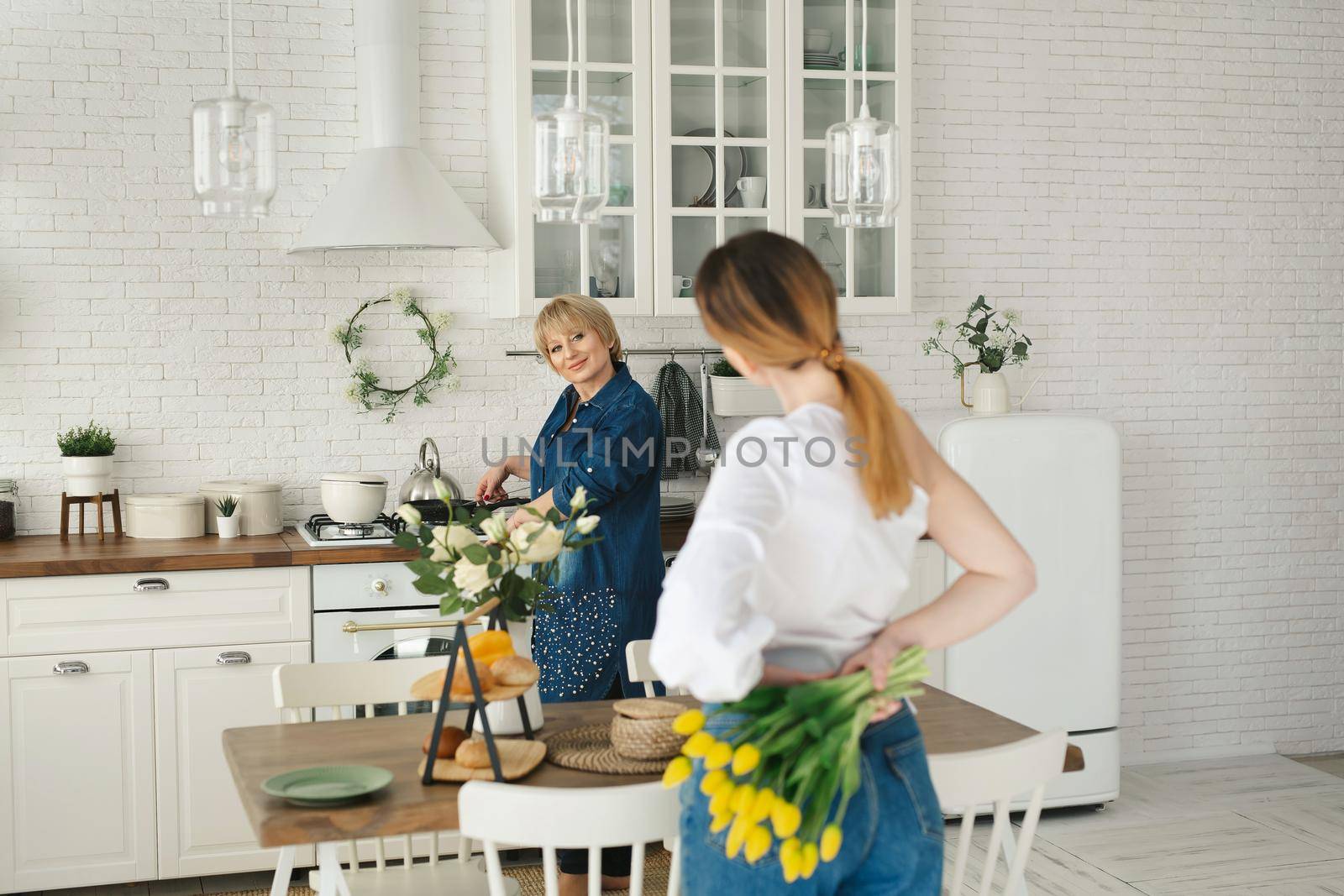 Beautiful adult daughter hides flowers behind her back for her adult mother. Mother's day, a women's holiday.