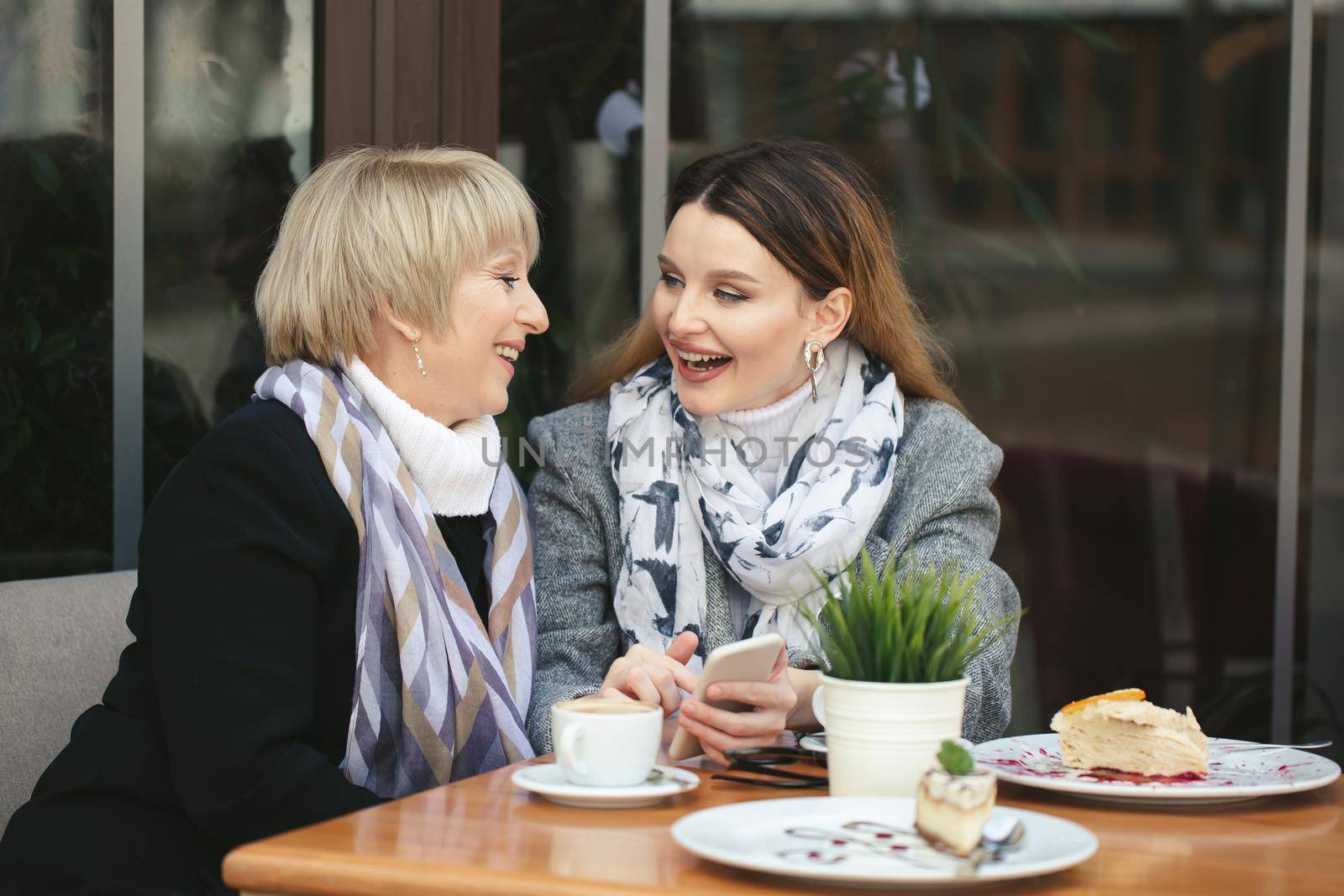 Family, an adult mother and daughter are sitting at a wooden table in a cafe on the street, looking at each other, smiling and holding hands by StudioPeace