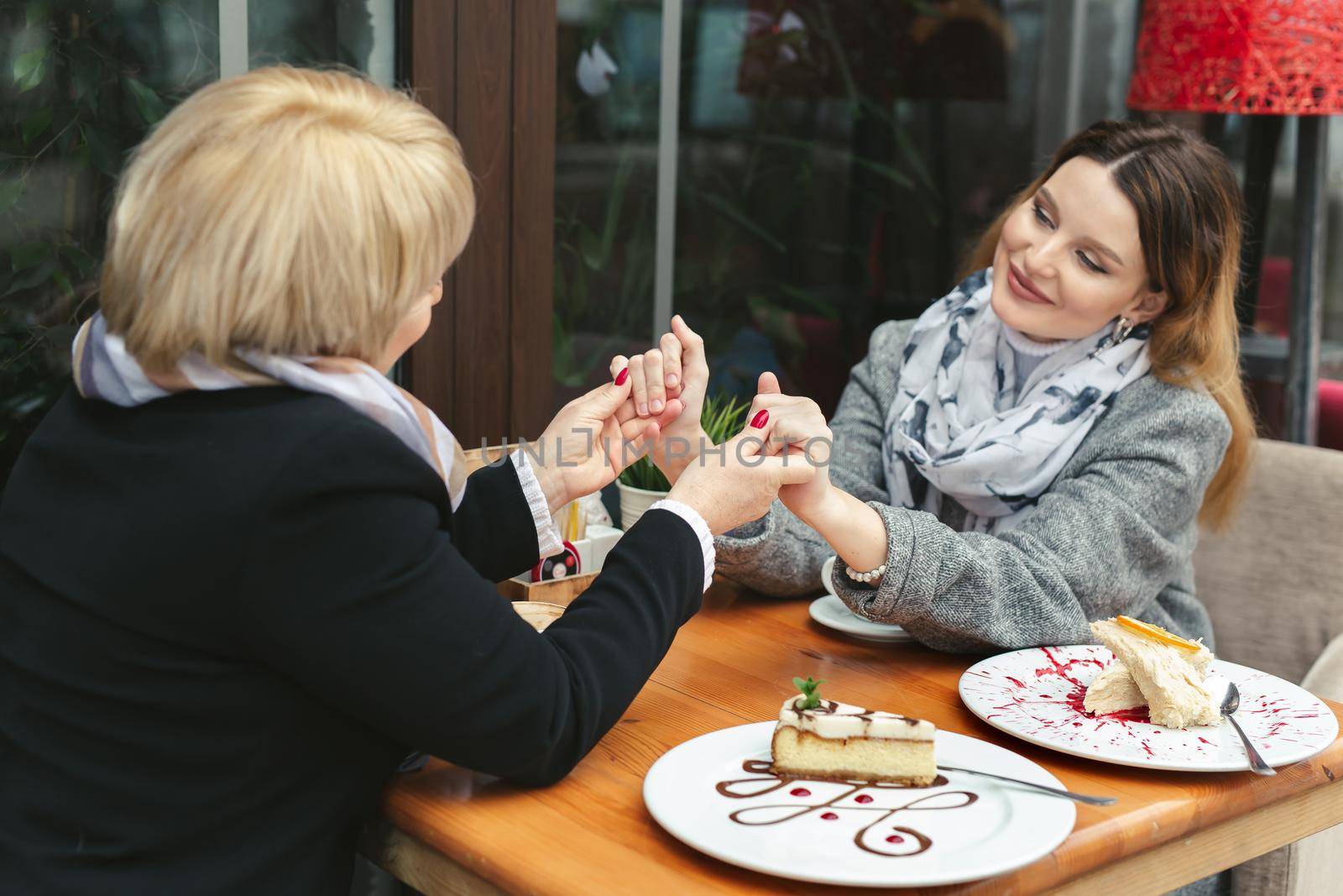 Family, an adult mother and daughter are sitting at a wooden table in a cafe on the street, looking at each other, smiling and holding hands by StudioPeace