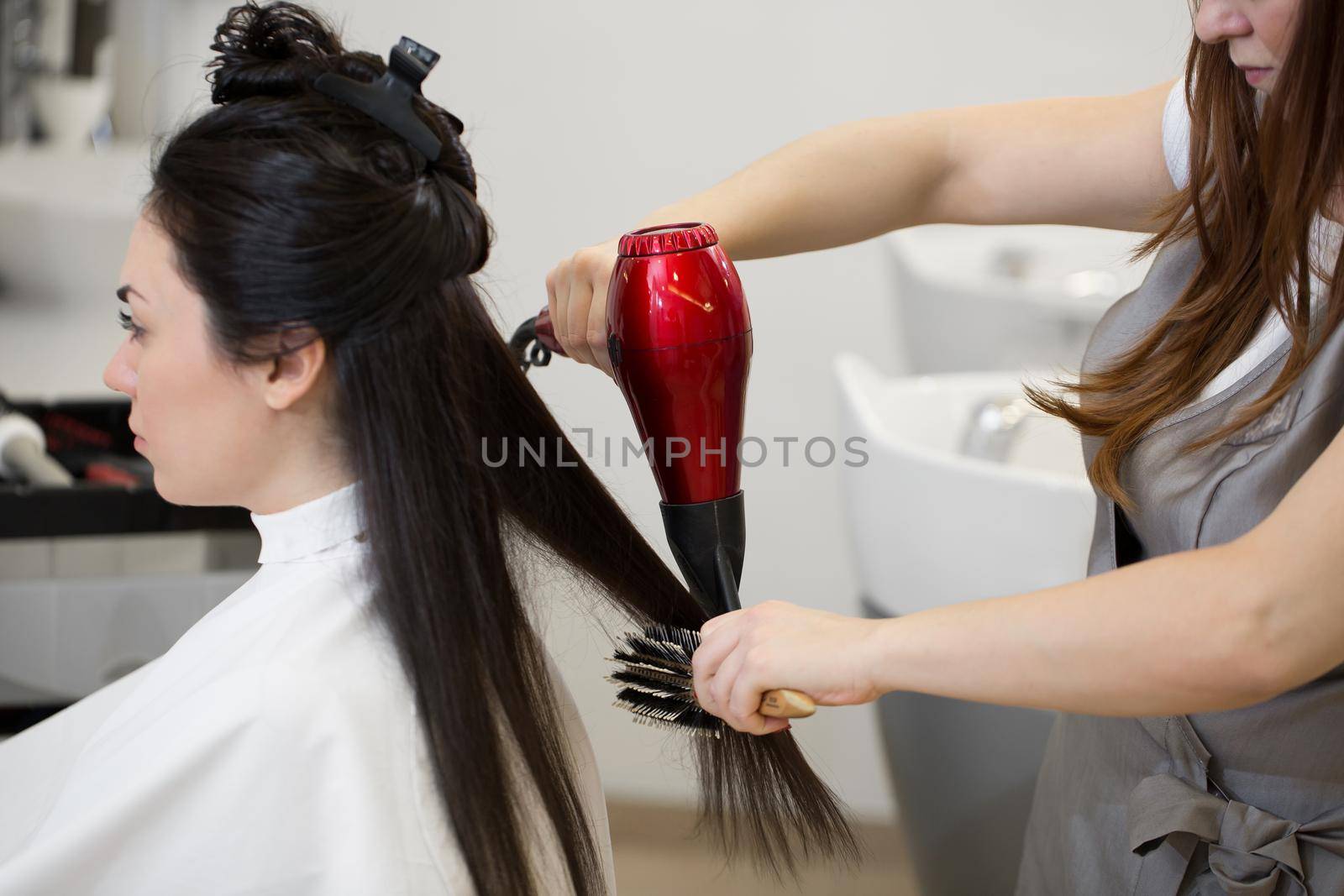 Hair stylist work on woman hairstyle in salon. Drying long brown hair with hair dryer and round brush